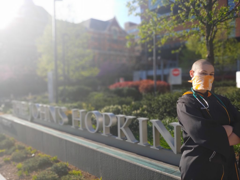 Bianca Palmisano in graduation gown and mask in front of a johns hopkins sign