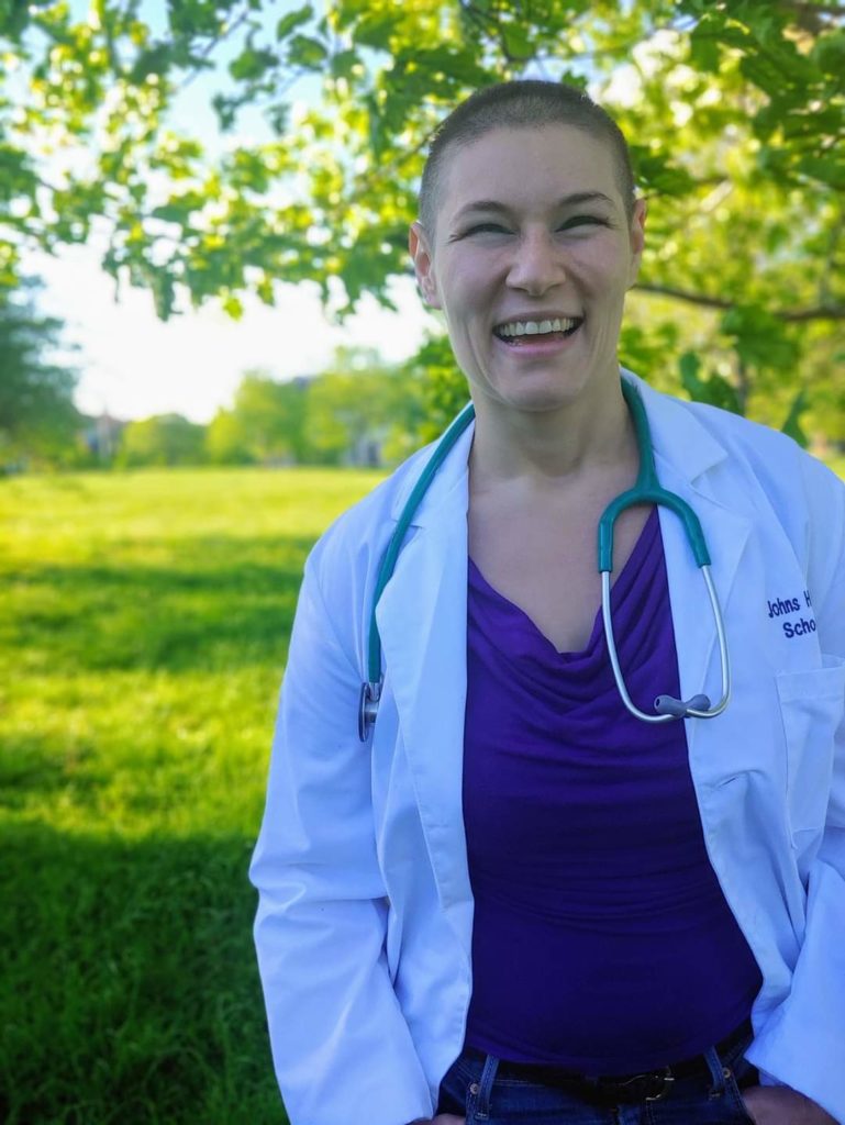 Bianca Palmisano smiles at the camera, in a white medical coat and a stethoscope around their neck